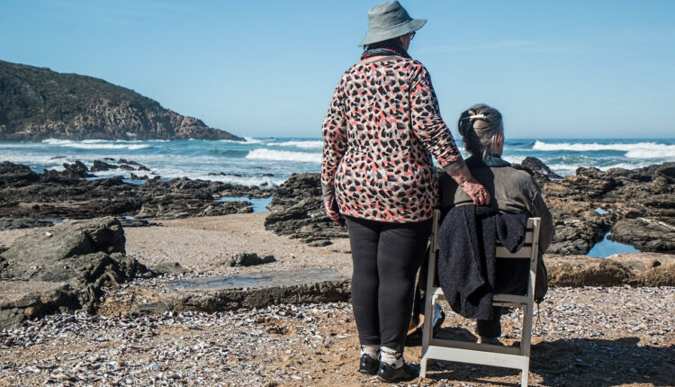 A person standing next to a person sitting in a chair on a beach Description automatically generated with medium confidence